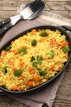 Tasty rice with vegetables in baking dish and cutlery on wooden table, top viewの写真素材
