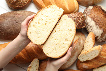 Woman with pieces of fresh bread over different other loafs at white table, top view