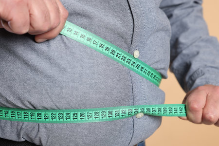 Overweight man measuring his belly with tape on beige background, closeupの写真素材