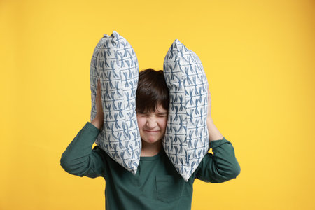 Annoyed boy covering his ears with pillows due to loud sound on yellow backgroundの写真素材