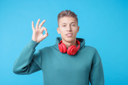 Young man with headphones showing OK gesture on light blue backgroundの写真素材