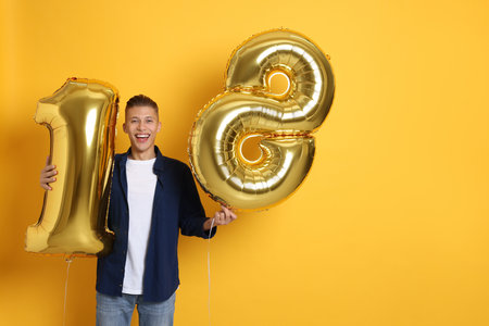 Coming of age party - 18th birthday. Happy young man with number shaped balloons on yellow background, space for textの写真素材