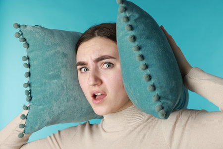 Distressed woman covering her ears with pillows from loud noise on blue backgroundの写真素材