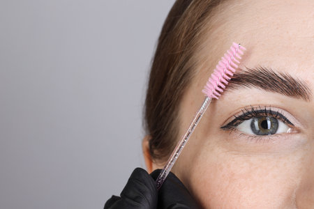 Brow lamination. Cosmetologist combing woman's eyebrows with brush against grey background, closeup....