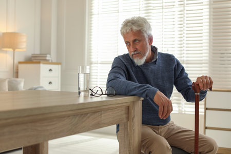 Lonely senior man with walking cane sitting at table in roomの写真素材