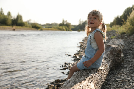 Cute little girl sitting on tree trunk near river. Child enjoying beautiful natureの写真素材