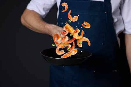 Chef cooking shrimps with chili pepper in frying pan on black background, closeupの写真素材