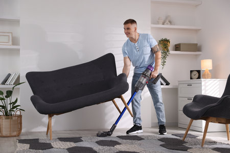 Young man cleaning rug under sofa with cordless vacuum cleaner at homeの写真素材