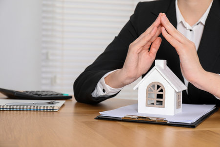 Property insurance. Real estate agent protecting house figure at wooden table indoors, closeupの写真素材