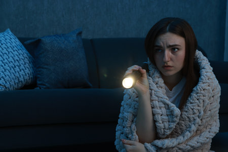 Fear of darkness. Scared young woman with flashlight and blanket near couch indoors at night, space for textの写真素材