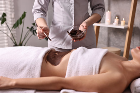 Chocolate body wrap. Spa worker applying mask onto woman's belly in salon, closeupの写真素材