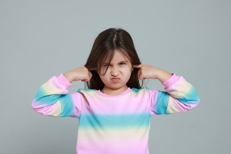Annoyed little girl covering her ears from loud sound on grey backgroundの写真素材