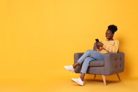 Smiling woman with smartphone sitting in armchair on yellow background. Space for textの写真素材