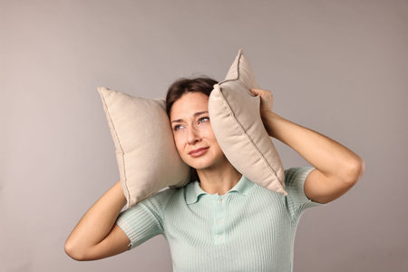 Annoyed woman covering her ears with pillows due to loud sound on light grey backgroundの写真素材