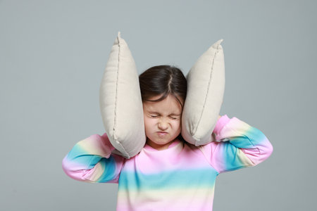 Annoyed little girl covering her ears with pillows from loud sound on grey backgroundの写真素材