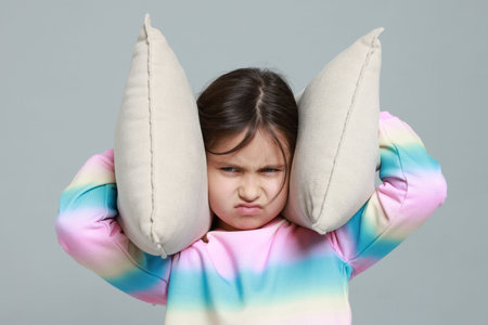 Annoyed little girl covering her ears with pillows from loud sound on grey backgroundの写真素材
