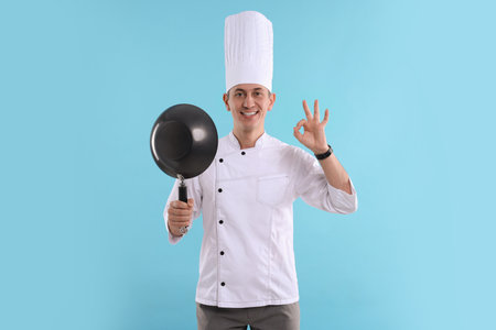 Professional chef with wok showing okay gesture on light blue background