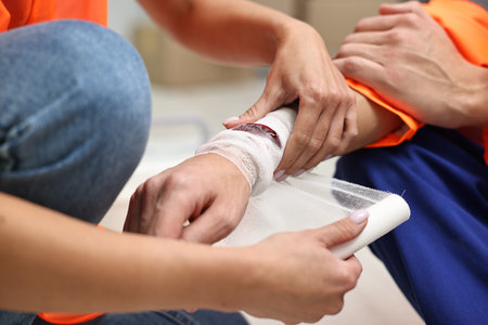 First aid. Woman putting bandage on her colleague's injured wrist after work accident indoors,...