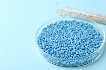 Colorful plastic granules in petri dish and test tube on light blue background, closeup. Space for...