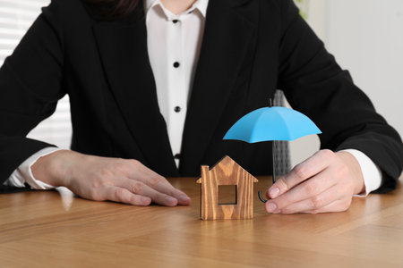 Property insurance. Real estate agent holding small umbrella above wooden house figure at table indoors, closeupの写真素材