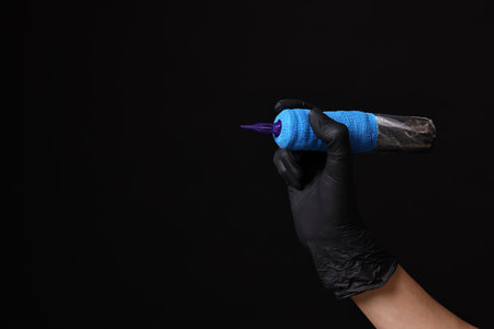 Professional artist in gloves holding tattoo machine on black background, closeup. Space for textの写真素材