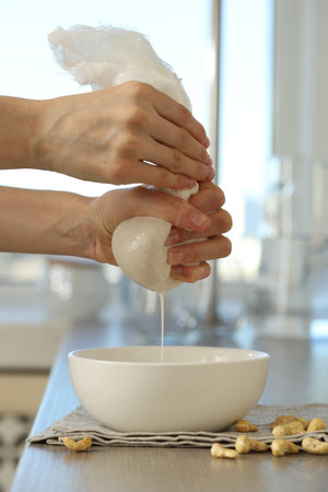 Woman straining cashew milk into bowl at wooden table with nuts indoors, closeupの写真素材