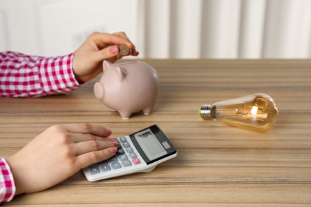 Woman putting coin into piggy bank while using calculator at wooden table, closeup. Energy saving...
