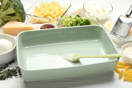 Ceramic casserole, brush and ingredients on white wooden table, closeup