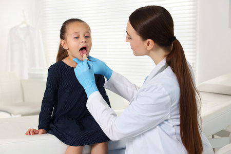 Doctor examining girl's throat with tongue depressor in clinicの写真素材