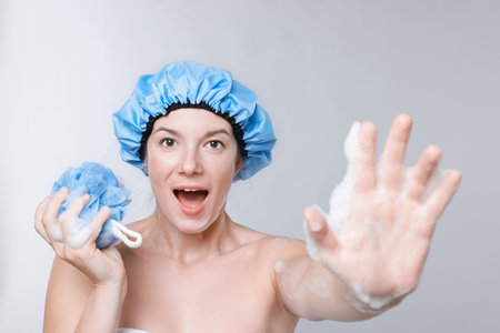 Emotional woman with shower cap and mesh sponge making stop gesture on gray backgroundの写真素材