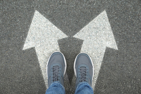 Man standing on asphalt road with white arrows pointing in different directions, top view. Concept of choice and making decisionsの写真素材
