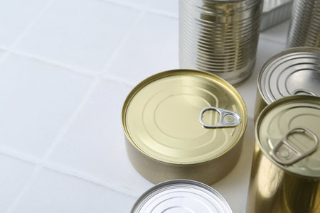 Many closed tin cans on white tiled table, closeup. Space for text