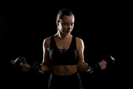 Woman in sportswear training with barbells on black background