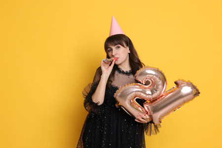 Coming of age party - 21st birthday. Young woman with hat and blower holding number shaped balloons on yellow backgroundの写真素材