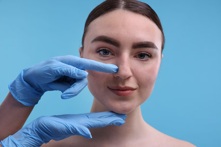 Doctor checking patient's nose before plastic surgery operation on light blue background, closeupの写真素材