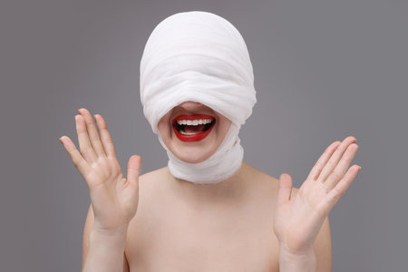 Woman with face wrapped in medical bandage after plastic surgery operation on gray backgroundの写真素材