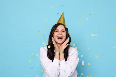 Happy woman in party hat under falling confetti on light blue background. Surprise partyの写真素材