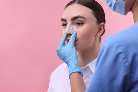 Doctor checking patient's nose after plastic surgery operation on pink background, closeup. Space for textの写真素材