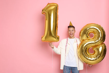 Coming of age party - 18th birthday. Happy young man with number shaped balloons and hat on pink...