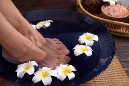Woman soaking her feet in bowl with water and plumeria flowers on floor, closeup. Spa treatmentの写真素材