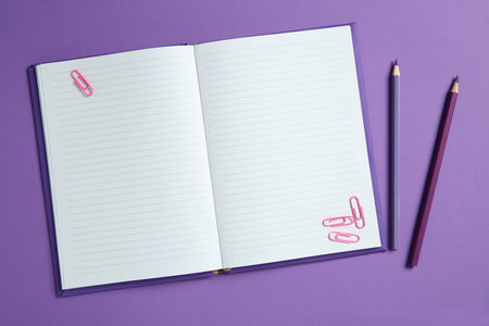 Blank notebook with paper clips and pencils on purple background, top view. Space for textの写真素材