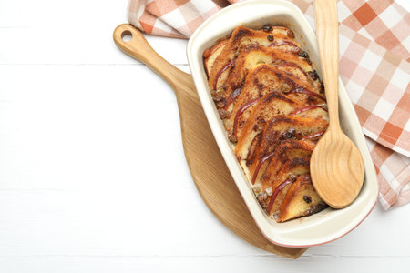 Delicious bread pudding in baking dish and spoon on white wooden table, top view. Space for textの写真素材