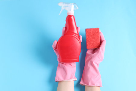 Woman with spray bottle of cleaning product and sponge on light blue background, top view. Space for...