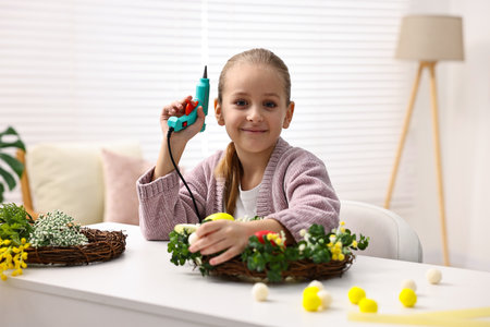 Little girl with glue gun creating Easter composition at table indoors