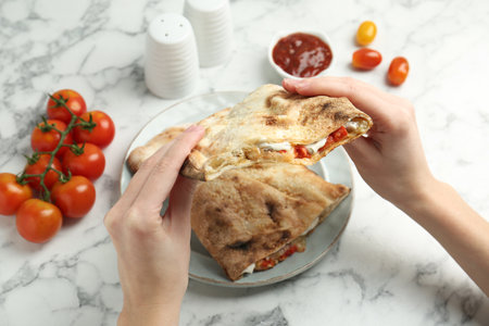 Woman eating tasty calzone with meat, cheese and tomato at white marble table, closeup