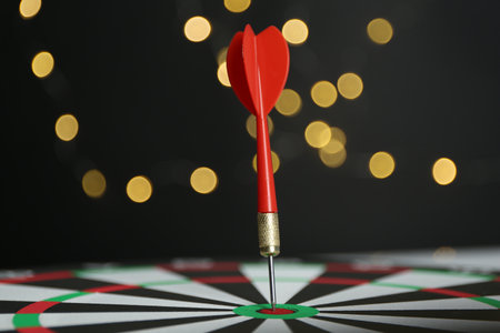 Red arrow hitting target on dart board against black background with blurred lights, closeupの写真素材