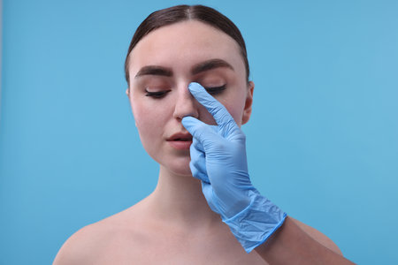 Doctor checking patient's nose before plastic surgery operation on light blue background, closeupの写真素材