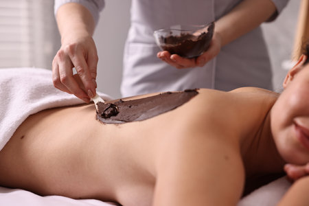 Chocolate body wrap. Spa worker applying mask onto woman's back in salon, closeupの写真素材