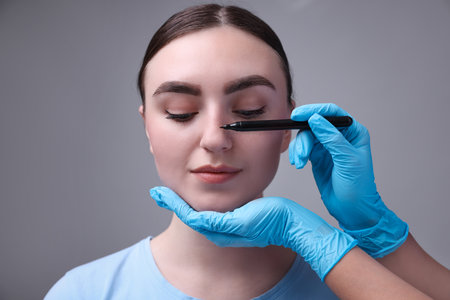 Doctor drawing marks on patient's nose for plastic surgery operation against grey background, closeupの写真素材