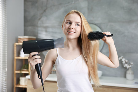 Beautiful young woman styling her hair with hairdryer and brush in bathroomの写真素材